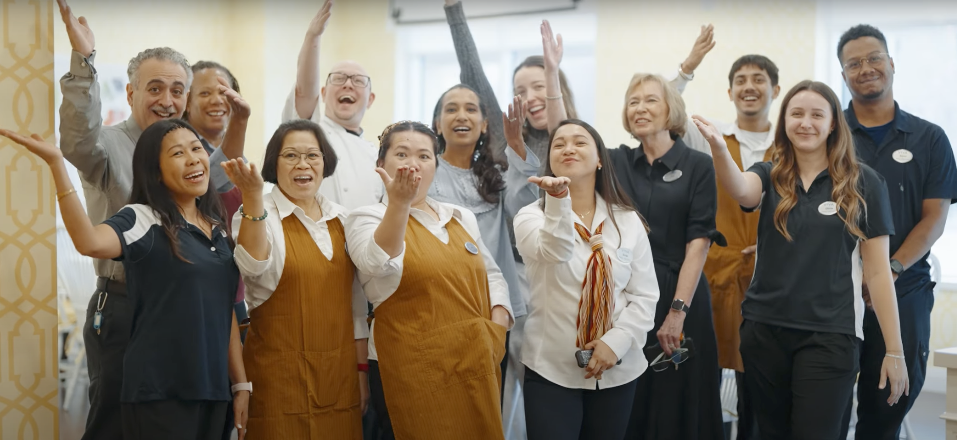 A group of senior living employees smiling for the camera and holding out hands in a welcoming way.